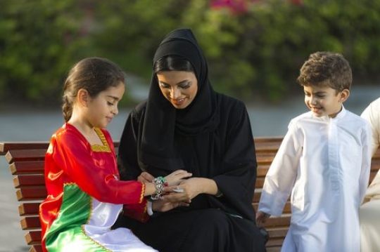Mother helping little girl with her bracelets while the son and father are sitting on the bench with them|-