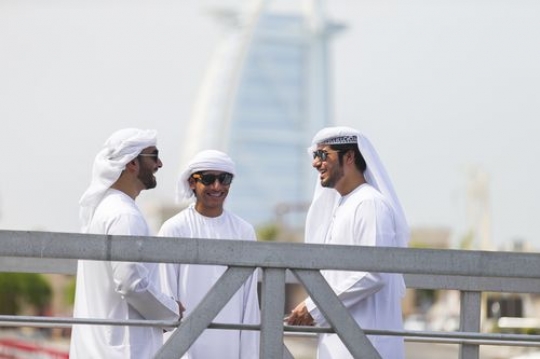 Men standing by the dock dressed in the traditional UAE attire talking to each other|